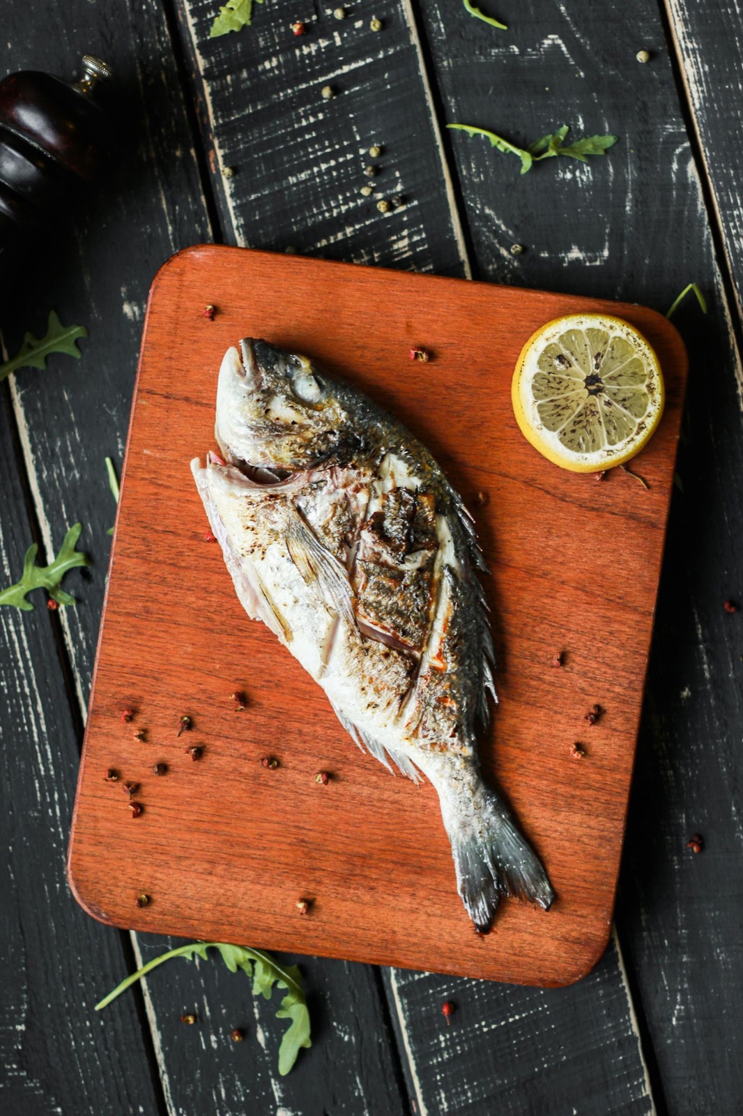 Whole grilled branzino on a wooden board with a slice of lemon and arugula.
