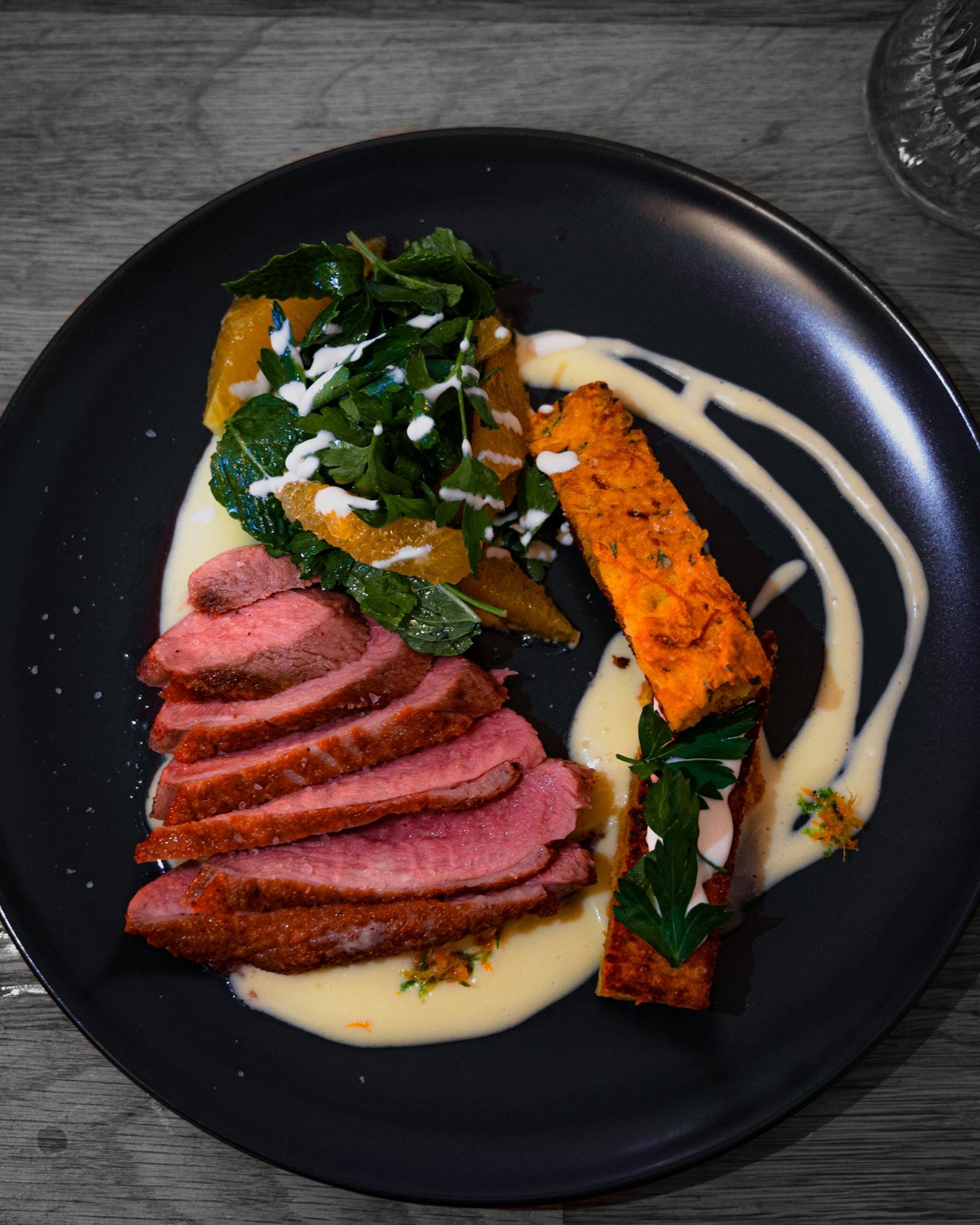 Fourteen-day dry-aged duck, sliced pink and plated with citrus and greens on a dark plate.